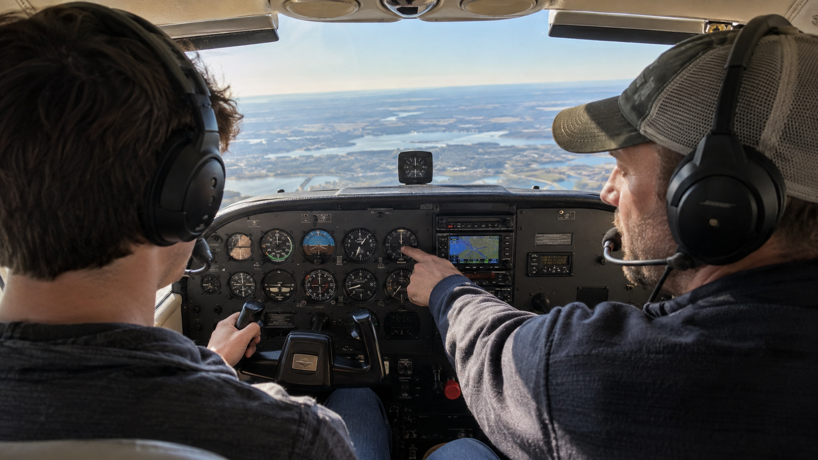 Two pilots wearing headsets in an aircraft cockpit during flight, with one pointing at instruments while scenic lakes and landscape visible through windscreen.